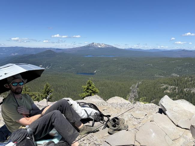 Man with umbrella relaxing on summit with mountain view in background