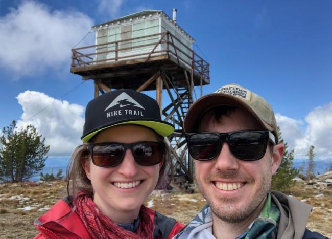 Man and woman take selfie in front of fire lookout tower. 
